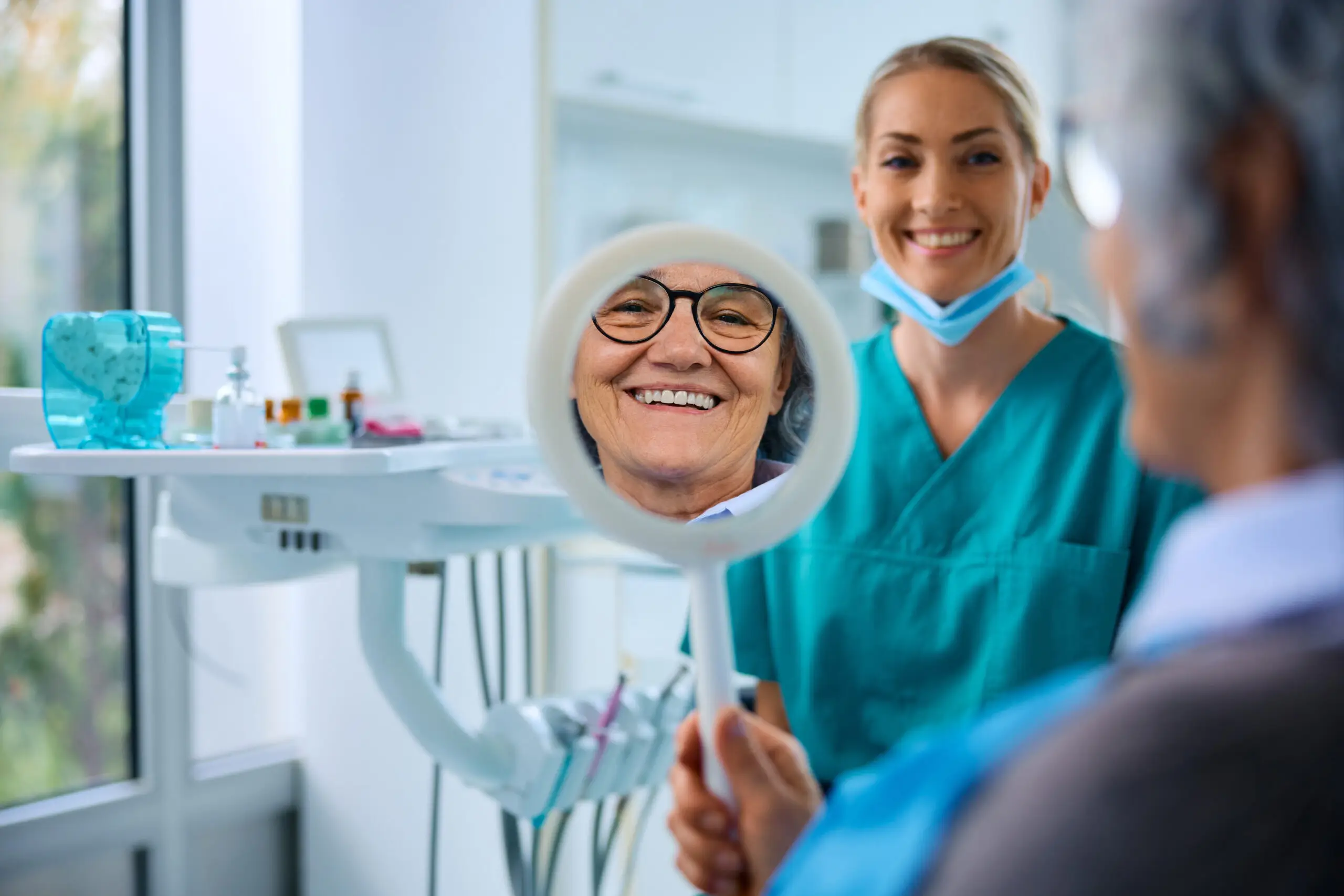 Smiling senior female patient in glasses looking in mirror at dental results with dentist.