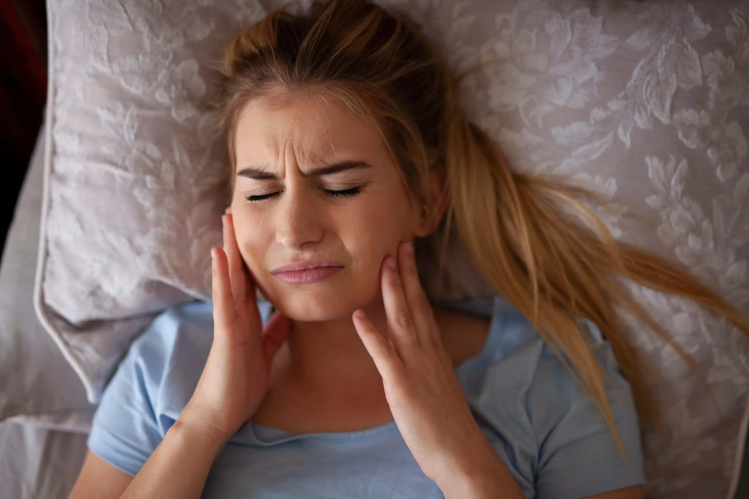 Young woman lying in bed holding her jaw with a pained expression due to teeth grinding.