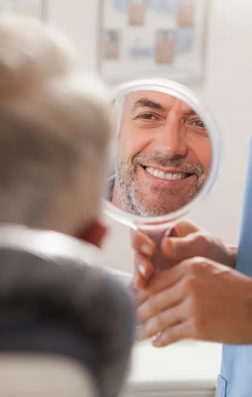 Man smiling and looking at his reflection in a handheld mirror held by a dental professional