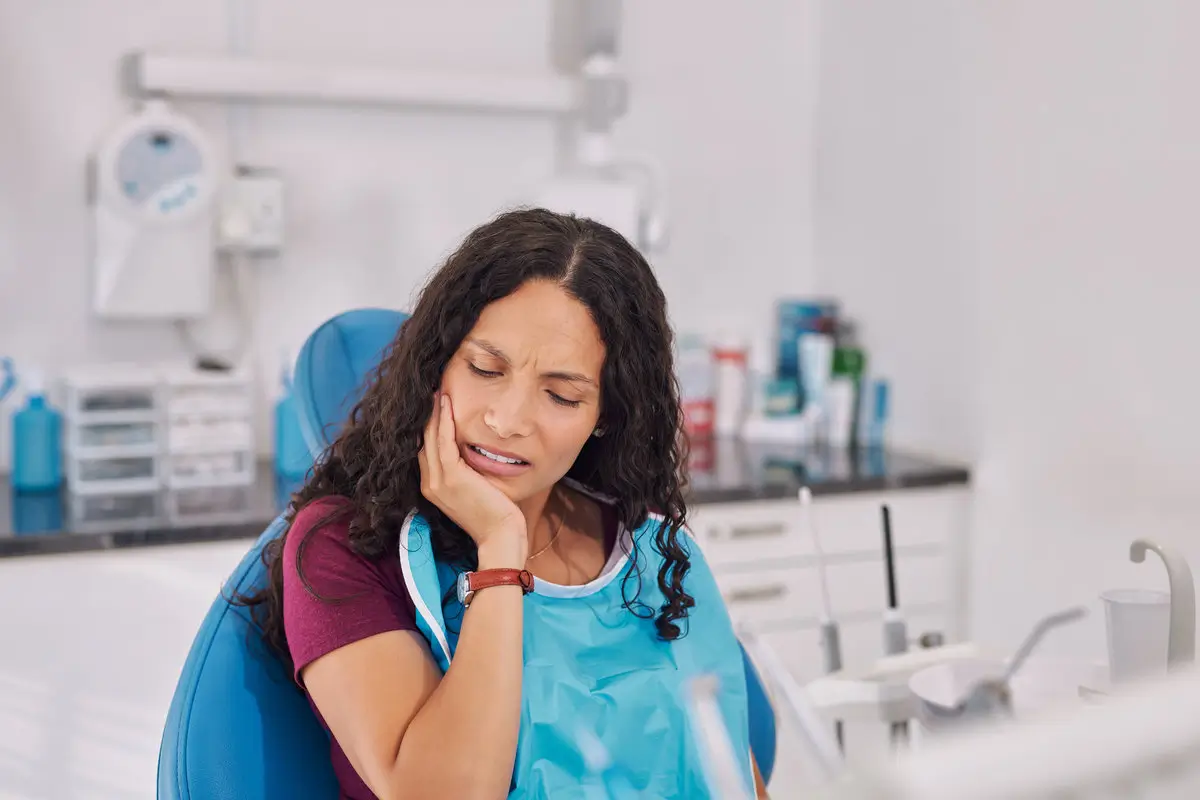 Young mixed race woman sitting in dental chair holding jaw in discomfort. Patient showing signs of tooth pain during consultation, oral infection or caria. Woman wearing dental bib in clinic with hands on face pressing her cheek, showing discomfort from a toothache.