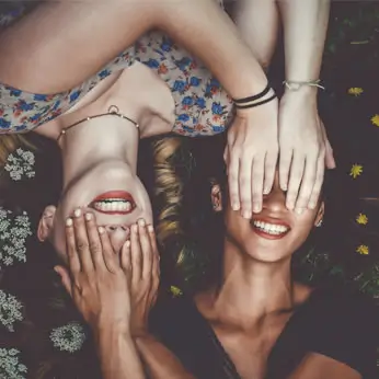 Two young women lying in grass, smiling and covering each other's eyes.